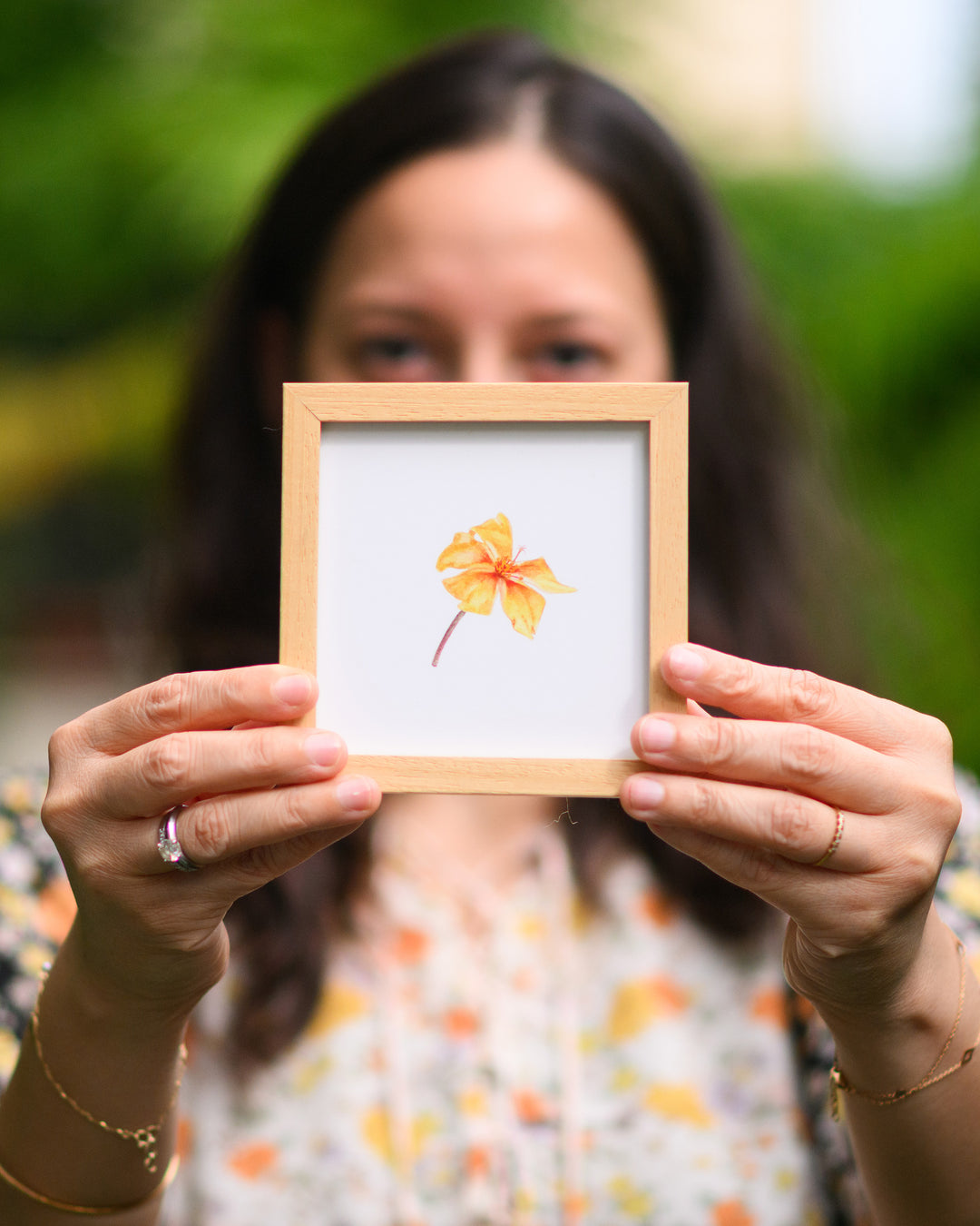 Person holding a small framed artwork of a yellow flower against a blurred green background