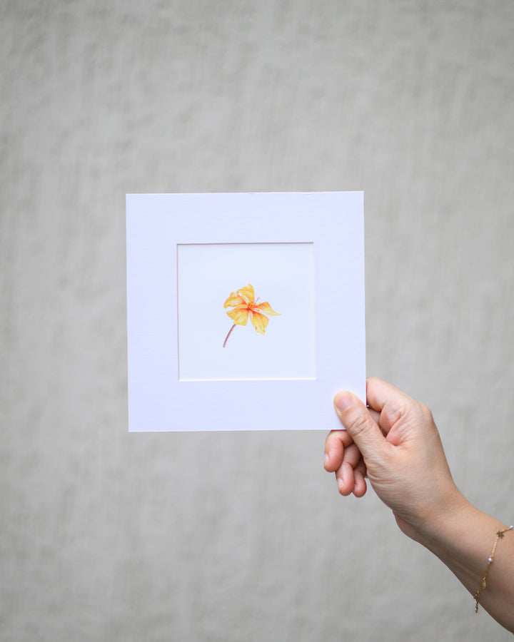 Hand holding a small watercolor painting of a flower on a light purple background against a gray background